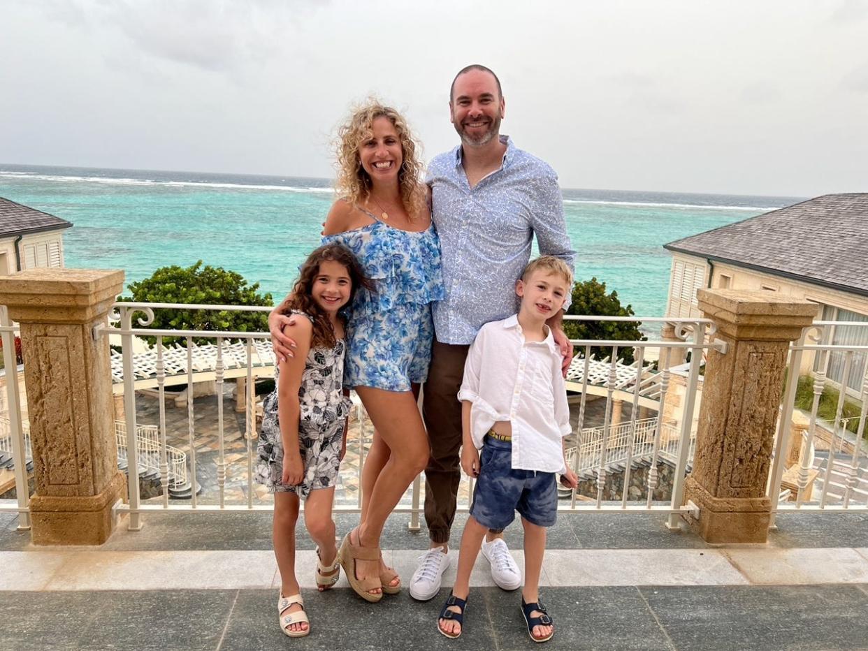 A family of four including a mother, father, and two young children pose in a tropical location.