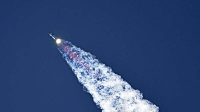 A SpaceX rocket flies across the frame from right to left, with engines bright at the bottom of the rocket, and smoke flying out of the bottom.