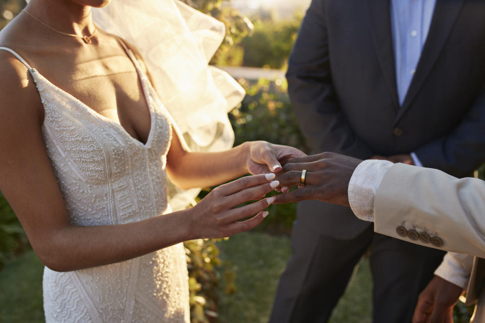 Bride in a lace wedding gown places a ring on the groom's finger while an officiant in a suit observes