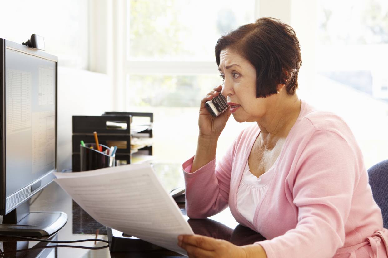 Side-view of a worried senior woman on her phone while looking at the desktop screen at her desk, holding a stack of papers, large open window showing trees outside in the background