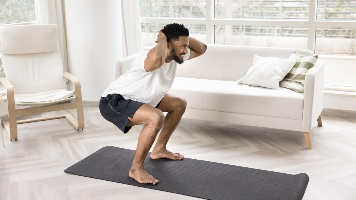  Man doing squat exercise on a yoga mat in pristine living room. 