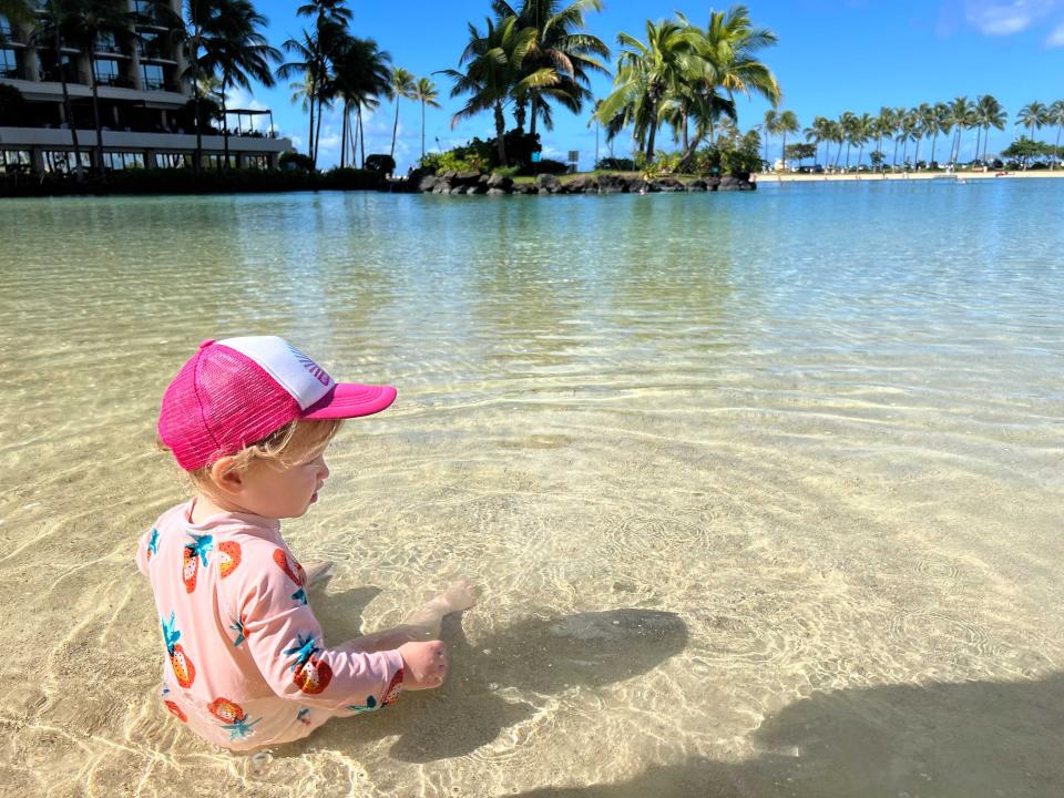 Little girl with a pink hat on sitting in a shallow lagoon with palm trees in the distance.