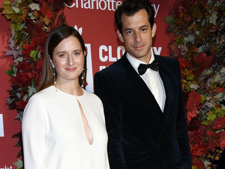 Grace Gummer, in a white gown, white handbag, and black heels, poses for photos with husband Mark Ronson, in a tuxedo, during an event.