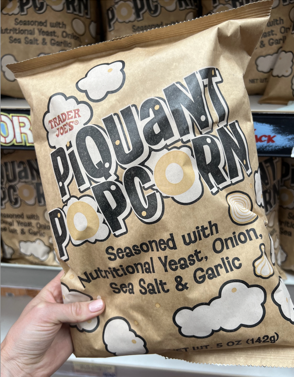 A hand holds a bag of Trader Joe's Piquant Popcorn, seasoned with nutritional yeast, onion, sea salt, and garlic. Shelves with more bags are in the background