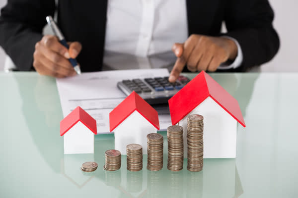 Close-up Of Businesswoman Calculating Invoice With Stacked Coins And House Models On Desk