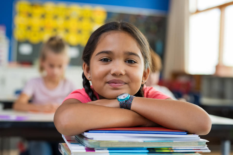 Portrait of cute little schoolgirl leaning on stacked books in classroom. Happy young latin girl in casual keeping chin on notebooks. Closeup face of smiling girl at elementary school.