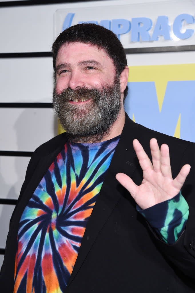 Man in a tie-dye shirt under a blazer, waving at the camera with a backdrop featuring logos