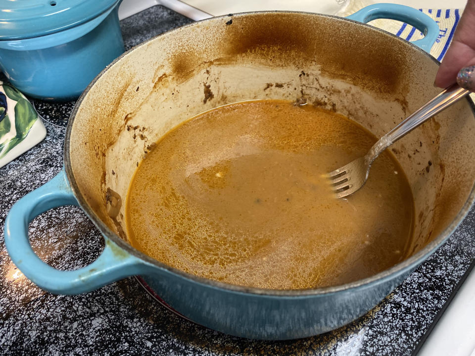 A partially filled blue pot on a stove with a fork stirring a light brown gravy or sauce