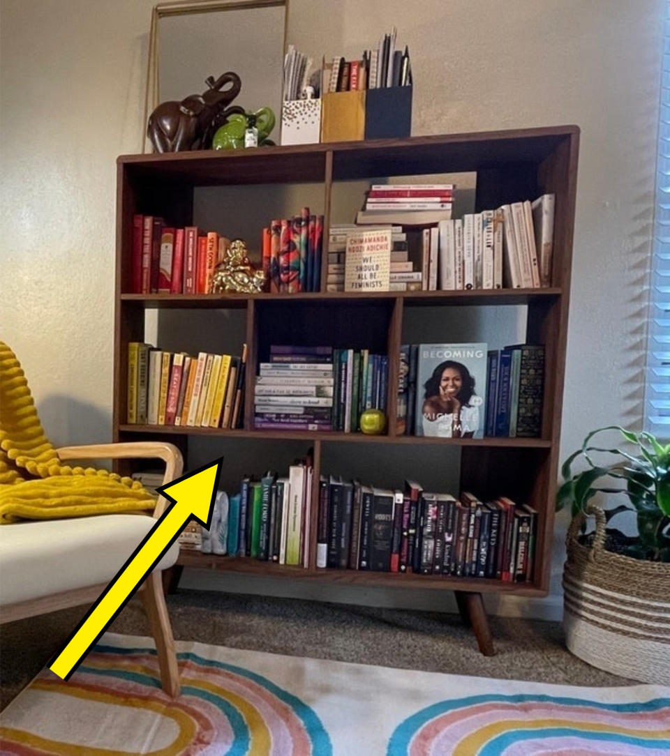 A cozy reading nook with a mid-century modern bookshelf filled with books and decor, a chair with a yellow throw, a plant basket, and colorful rug