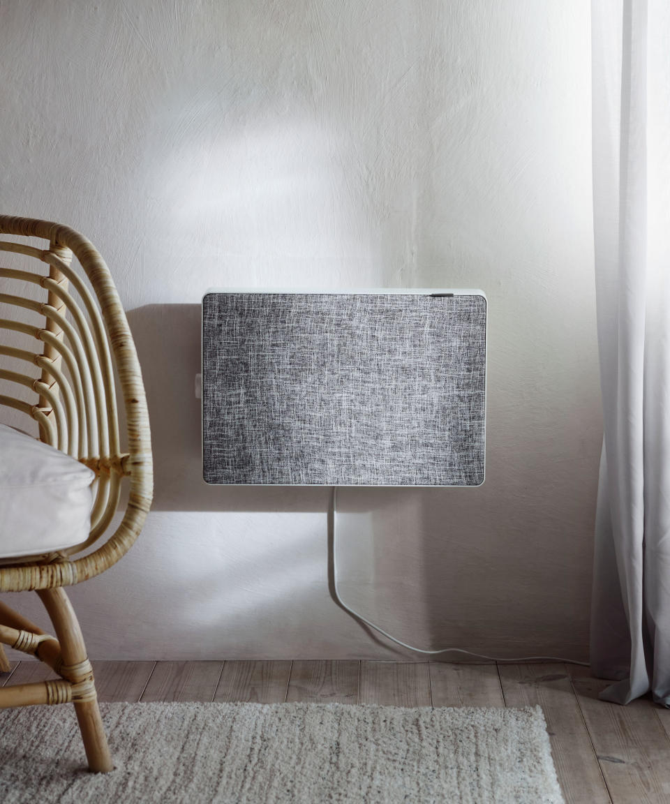 Gray IKEA air purifier mounted on the wall. there is a light wooden floor with mottled cream rug and wicker chair with cream seat cushion in view