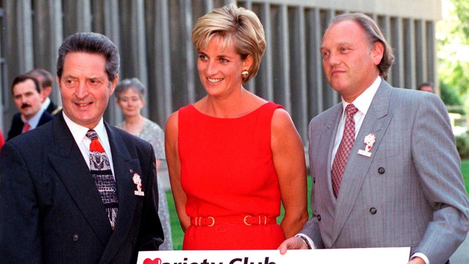 Princess Diana in a red scoop neck dress stood between two men in suits as they present her with a check thats being donated to Northwick Park Hospital