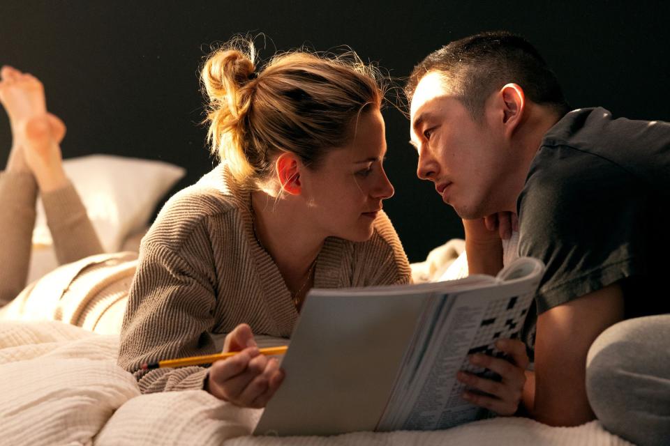 A still shows a cozy scene of Kristen Stewart and Steven Yeun in bed. She holds a pencil and a crossword book in her hands as he leans in for a kiss.