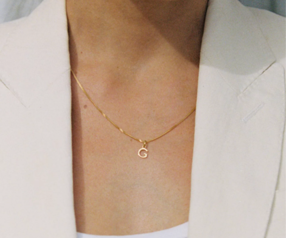 A gold chain with a red letter G lays on the chest of a woman in a cream blazer.