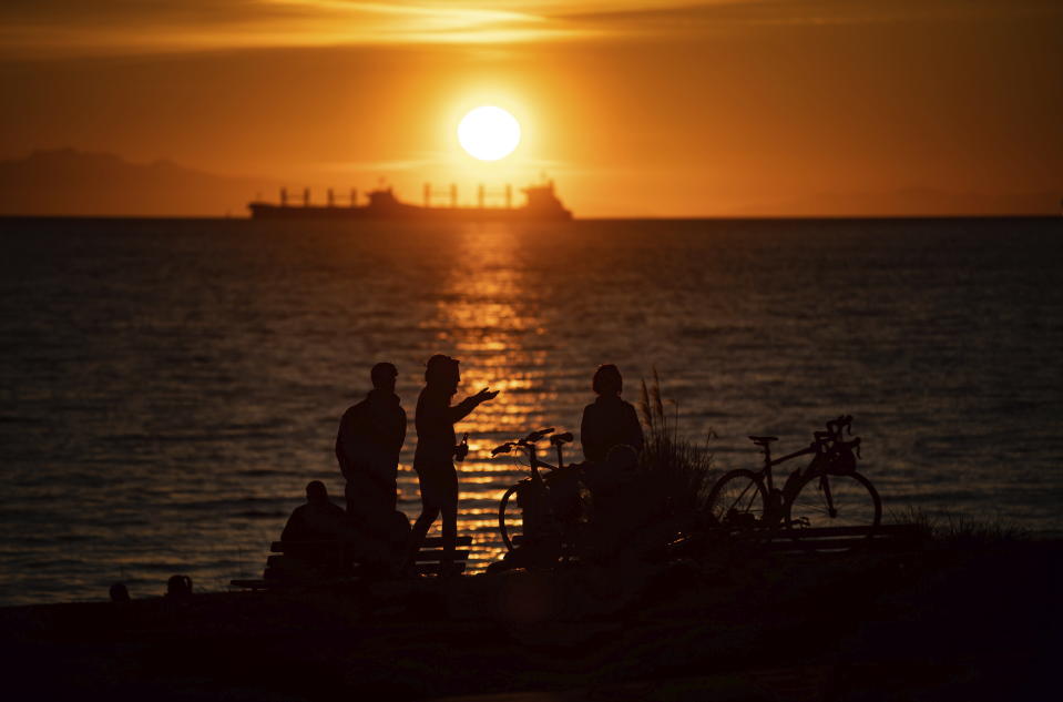 A woman is silhouetted while taking in the sunset with others at English Bay Beach, as a freighter is anchored in the distance off Vancouver, British Columbia, Saturday, March 21, 2020. The City of Vancouver asked those coming to the park and beach to maintain a distance of two meters between one another due to concerns about the spread of the coronavirus. (Darryl Dyck/The Canadian Press via AP)