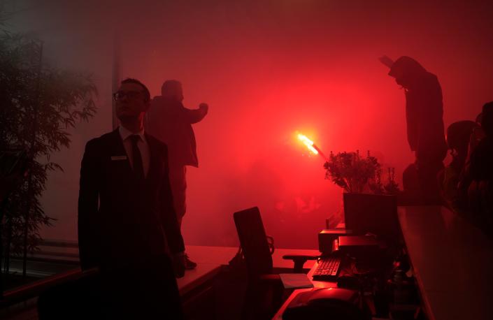 A man in a suit looking to the side while protestors with flares enter the LVMH building.