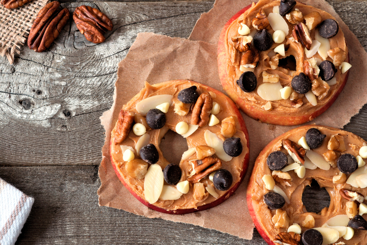 Three slices of apple with peanut butter and granola on a piece of paper bag on a rustic grey wooden table, surrounded by ingredients and napkins