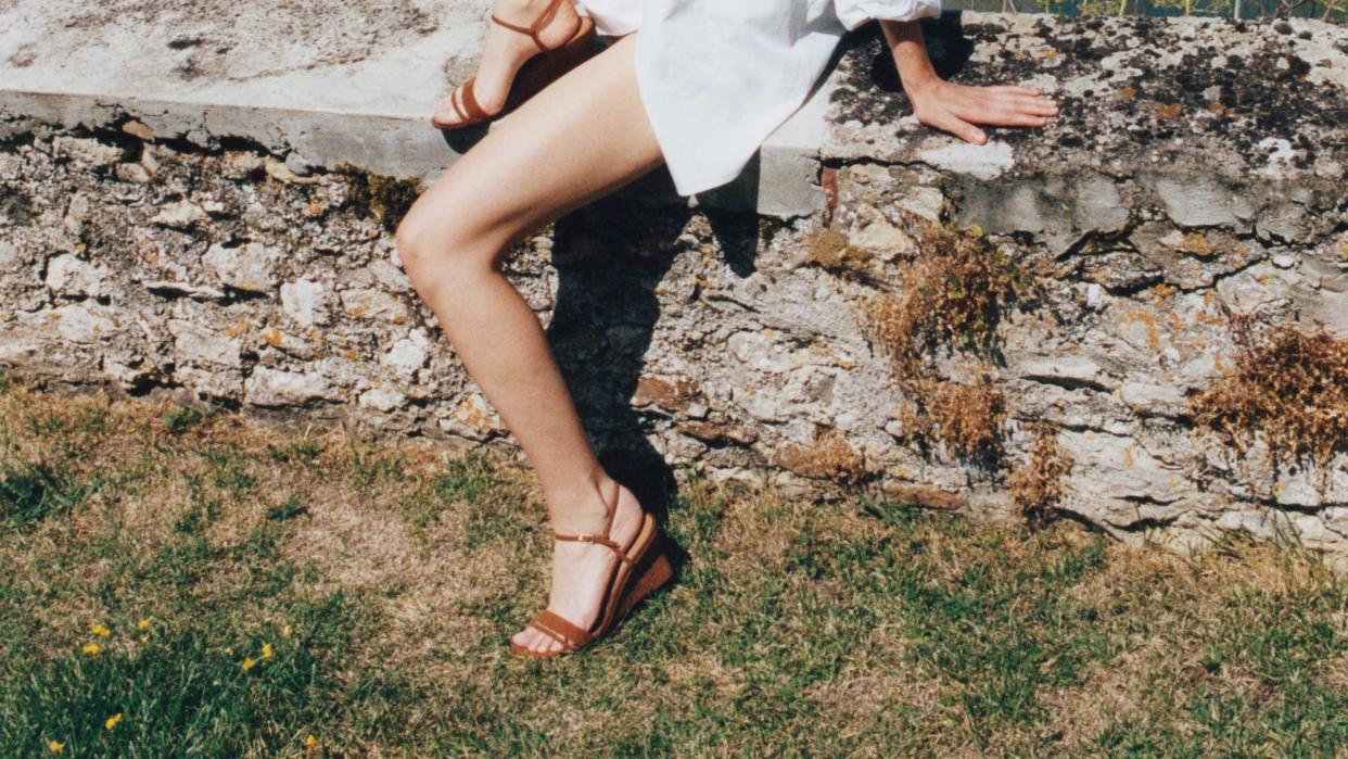 a model wears a pair of wedge sandals on a stone wall in a field