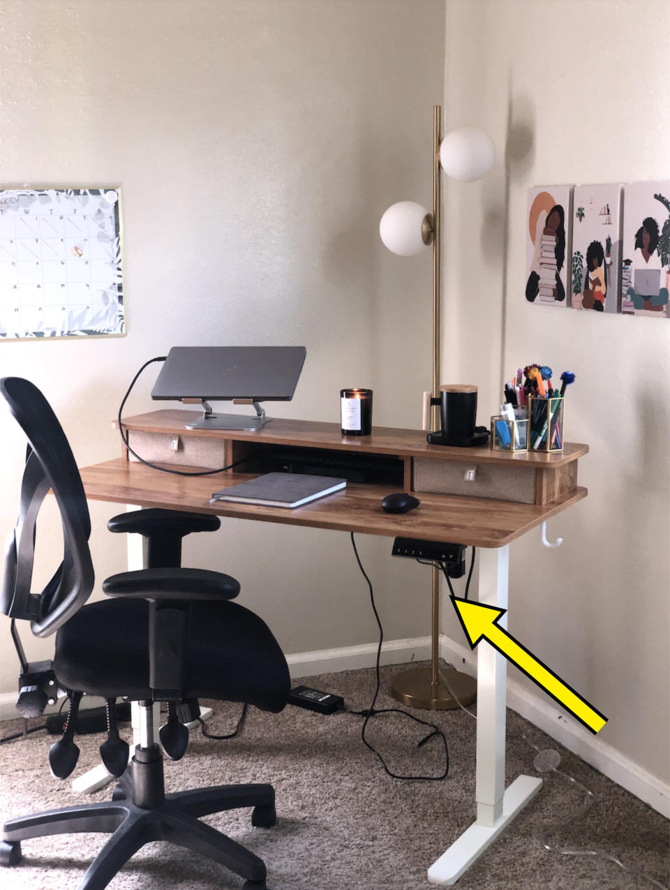 A well-organized home office setup with a wooden desk, ergonomic chair, lamp, and wall art. The desk has a laptop stand, pen holders, and a candle