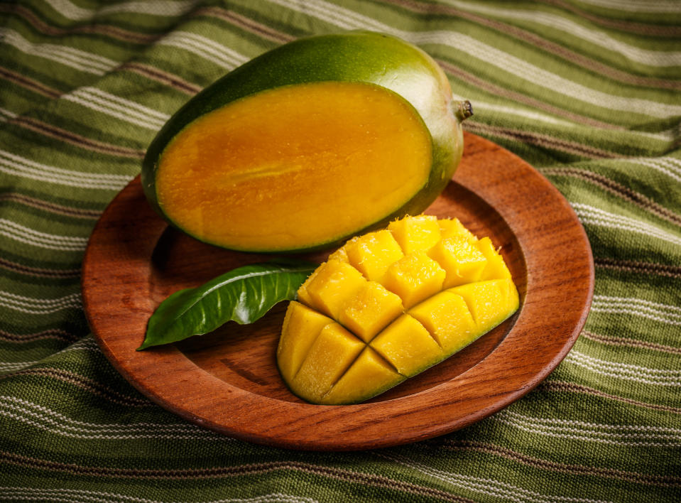 A mango and a green leaf are on a wooden plate. The mango has been sliced open with part of it cut into cubes. Underneath the wooden plate is a green towel with thin dark-green, white, and reddish-brown stripes