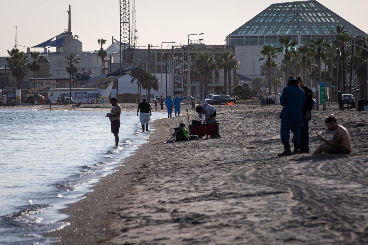 Men in blue suits walk the shoreline past beachgoers at North Beach during a cleanup effort by Flint Hills Resources on Jan. 3, 2023, in Corpus Christi, Texas. Ten days prior, an oil spill occurred originating from a pipe failure at Flint Hills Resources' Ingleside crude oil terminal.