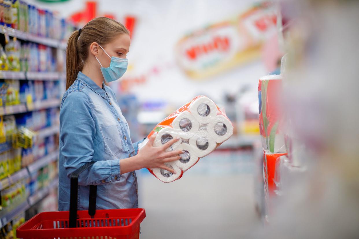 Young woman wearing protective face mask with shopping basket, holding pack of toilet paper and looking at it, COVID-19 pandemic era
