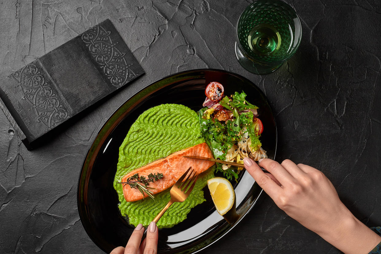 Top view of female hands cutting delicious grilled salmon steak served on plate with mashed peas, salad of fresh vegetables and lemon slice on black textured background