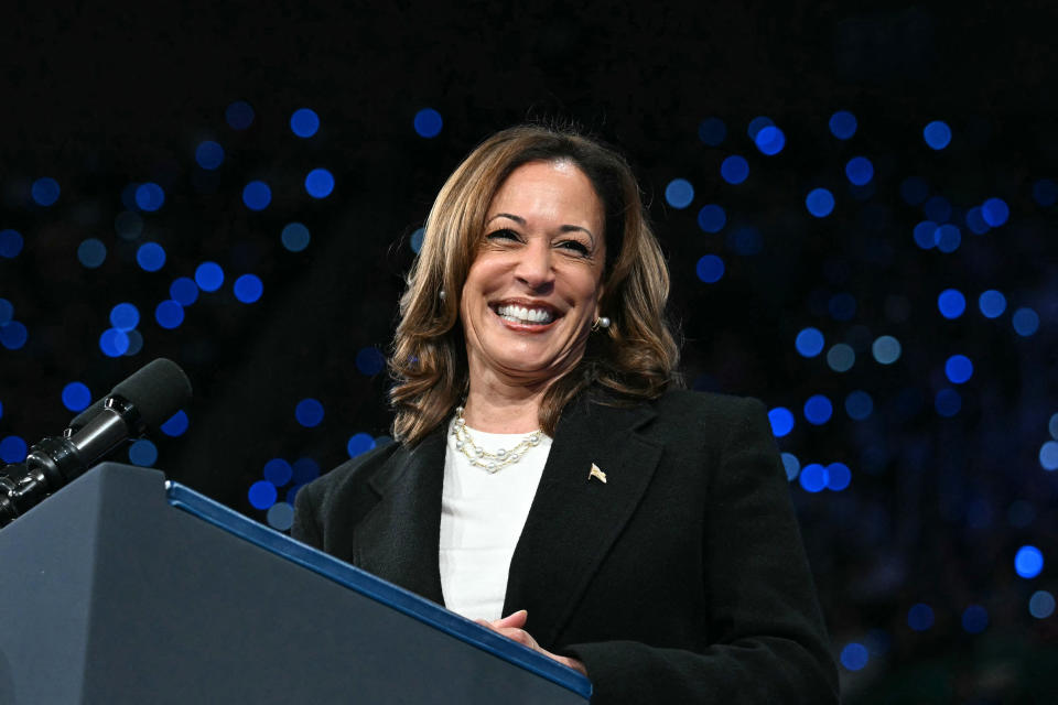 Kamala Harris smiles while speaking at a podium, with lights in the background.