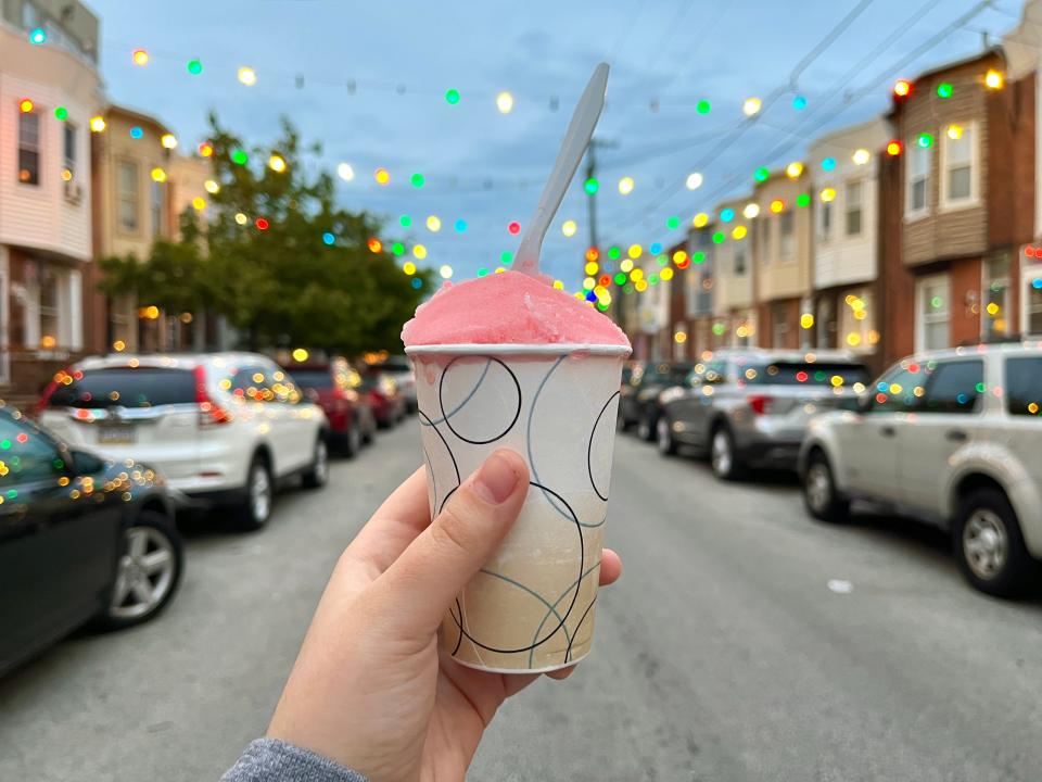 A hand holding a cup of pink water ice in front of a street with lights