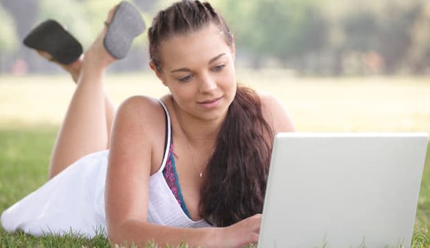 attractive young student working on lap top while lying on grass in park