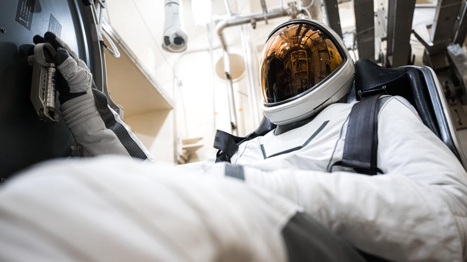     A figure in a clean white spacesuit sits strapped into a black bucket seat, with the image looking up from the blurred knee in the left foreground. Above left, a gloved hand with black-tipped fingers holds a component against the dark wall of a spacecraft. The full-face visor near the top center right of the helmet reflects a copper glow. Behind, white walls and pipes. 