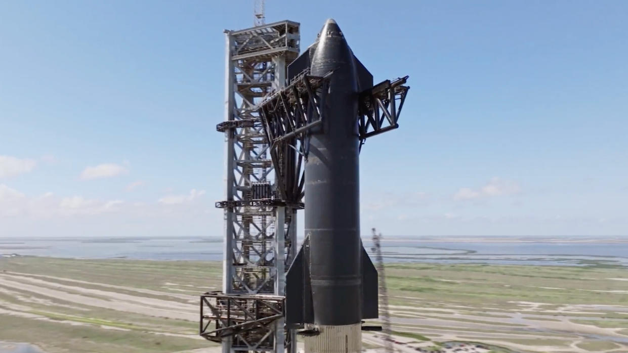  Closeup of a black, cylindrical spacecraft next to its gray launch tower, with greenish-brown wetlands and the sea in the background. 