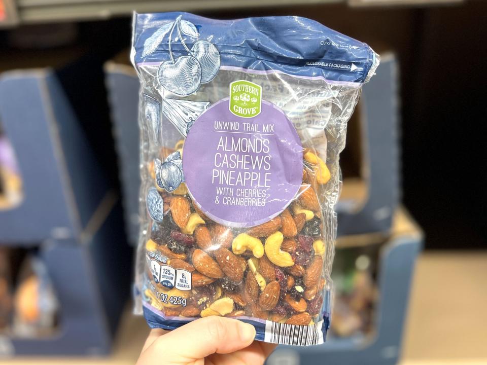 A hand holding a bag of Southern Grove trail mix, containing almonds, cashews, pineapple, cherries, and cranberries.