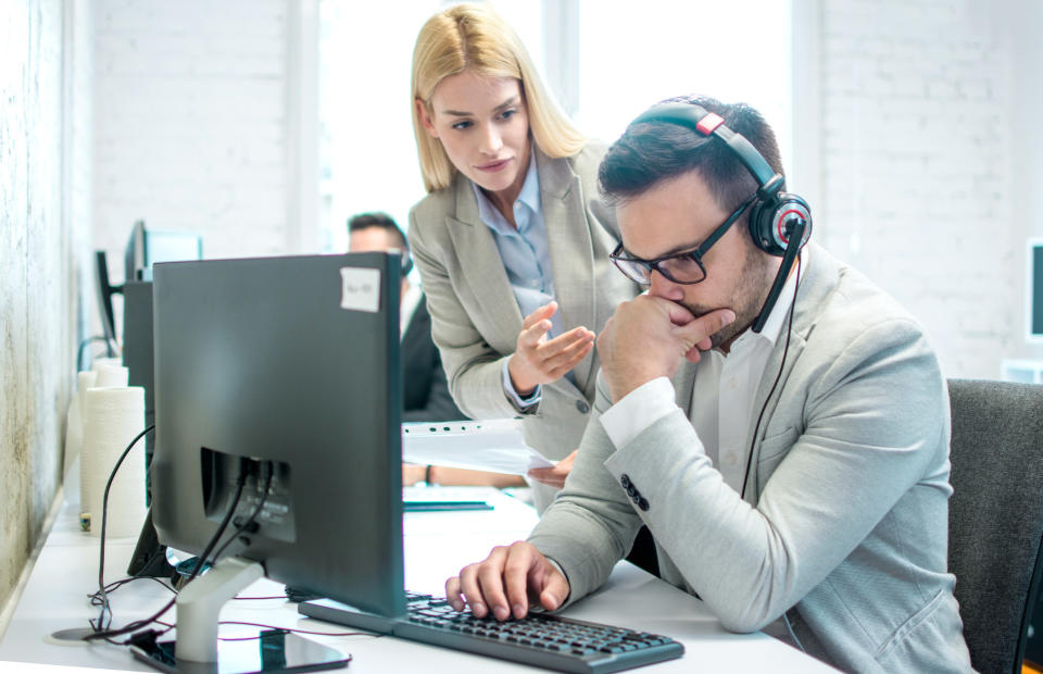 Woman standing over a seated man with headphones looking at a computer screen