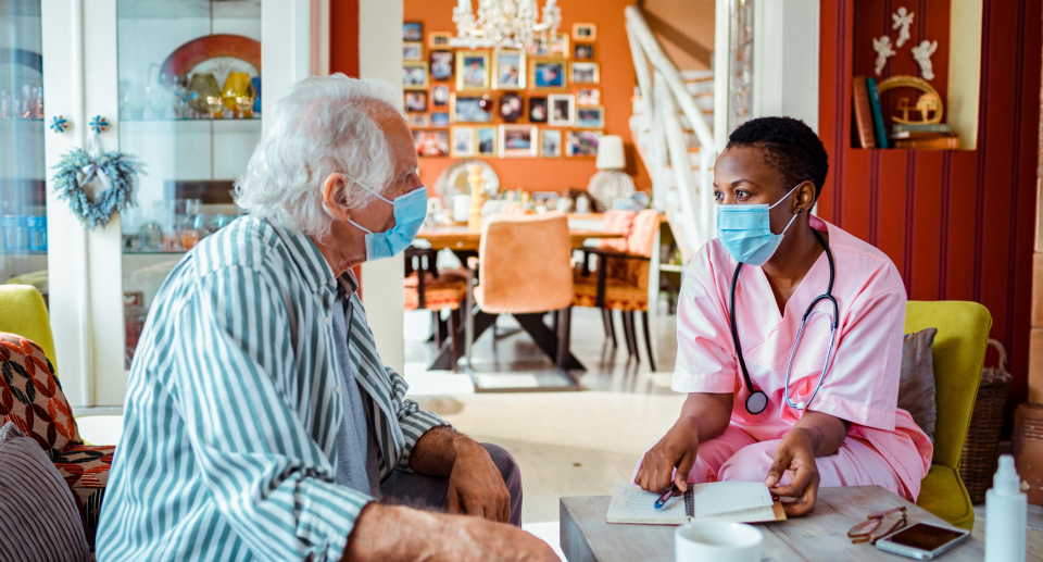 elderly man in pajamas sitting with nurse in pink scrubs wearing covid-19 face masks, In several parts of Canada, masks remain mandatory in healthcare settings, long-term care and retirement homes. (Photo via Getty)