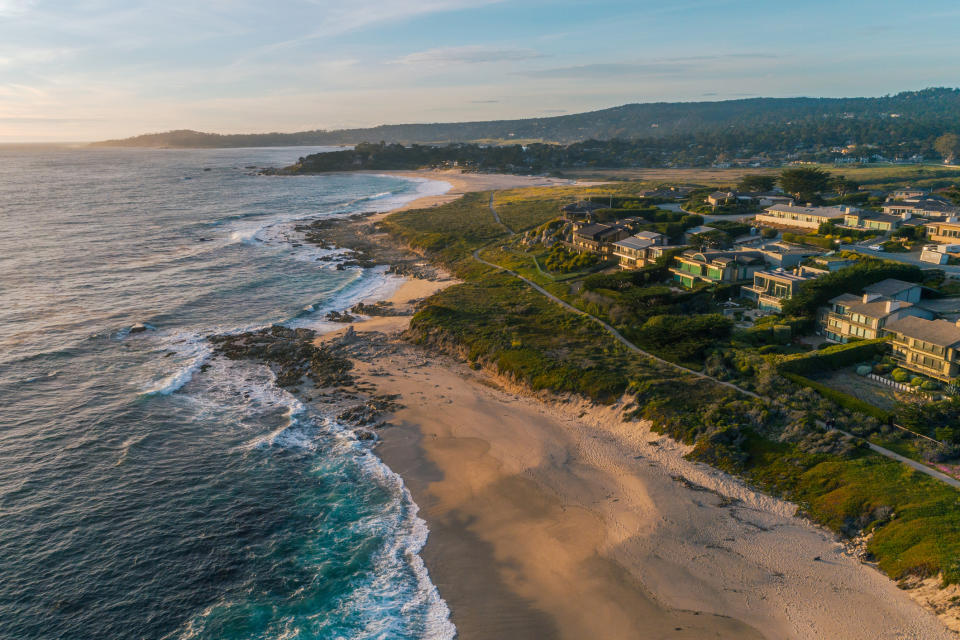 Aerial view of a coastal community with houses overlooking a beach and waves crashing on rocks, along with a scenic landscape in the background