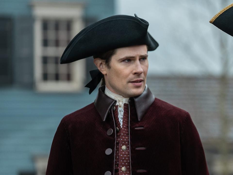 Lord John Grey wearing a black three-peaked hat and a dark red overcoat while talking to someone on his right.