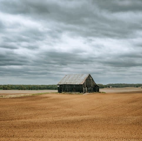 An old, weathered barn in a vast, plowed field under a cloudy sky