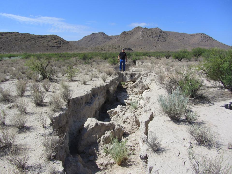 A ground fissure splits the desert ground heading towards a distant mountain range, with a surveyor standing on the edge in the distance. near Sulphur Hills (southeast of Willcox Playa