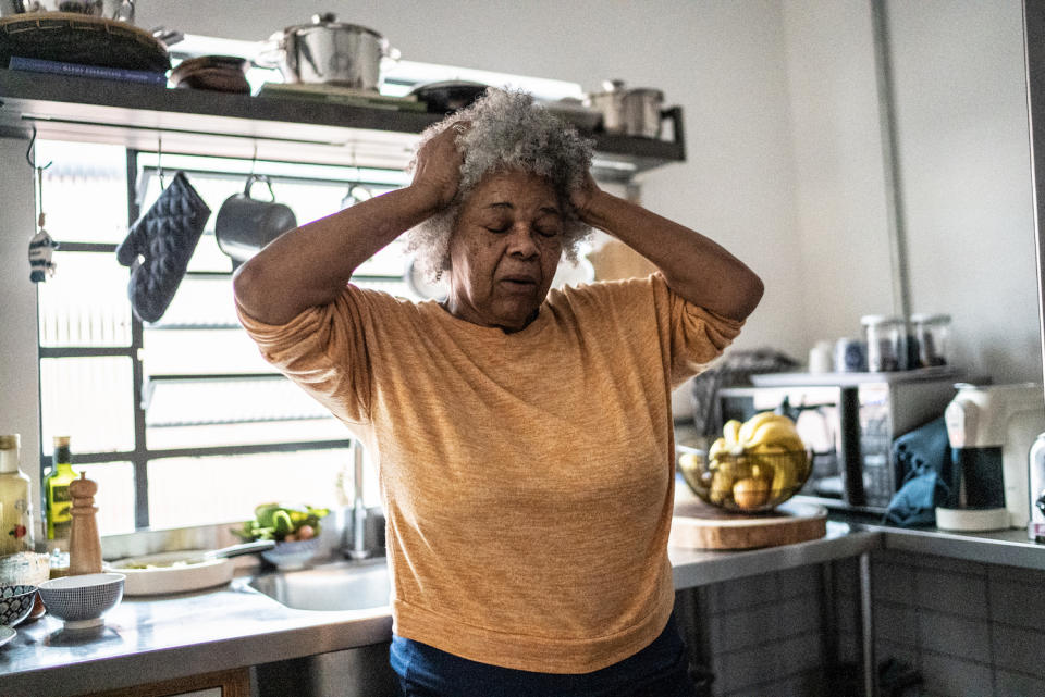 An older woman with her hands on the side of her head and her eyes closed while standing in her kitchen