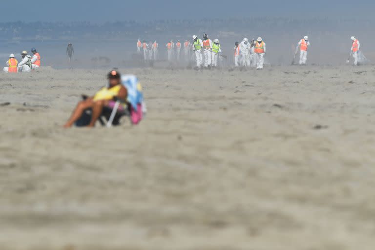 Un mujer toma sol en la playa mientras los equipos ambientales usan PPE mientras limpian los escombros después de un derrame de petróleo en el Océano Pacífico en Huntington Beach, California, el 5 de octubre de 2021