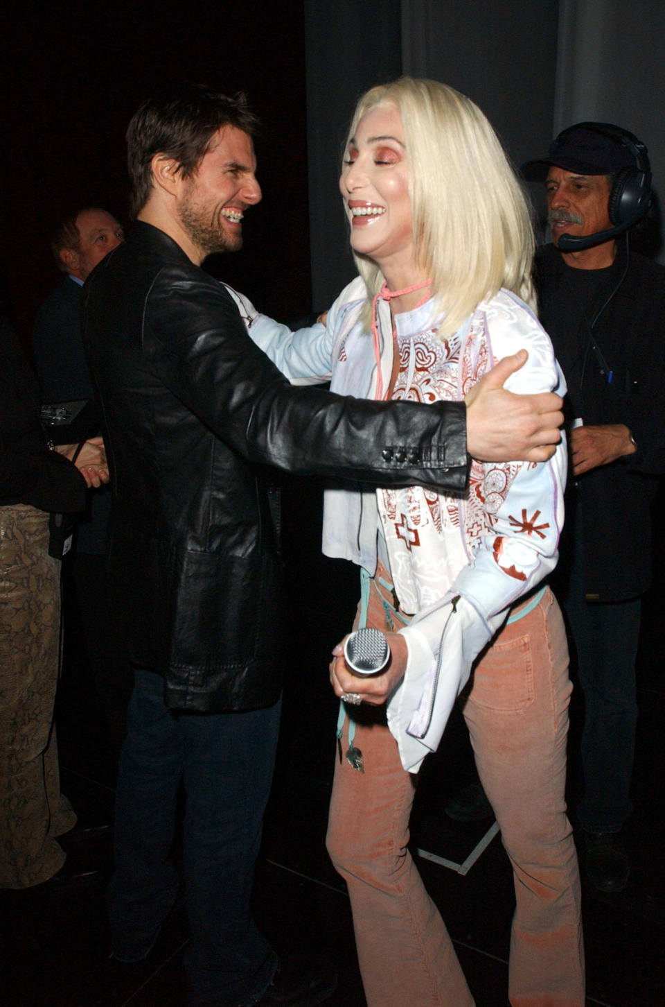 Tom Cruise and Cher in a half-embrace as she walks past him backstage, she's holding a microphone and they're both smiling