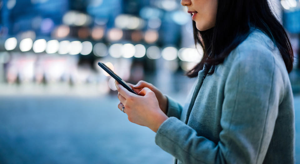 Close up of young Asian businesswoman using smartphone while commuting in downtown city street, with illuminated city street lights in background