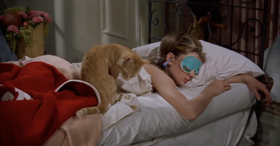 Audrey Hepburn lies in bed wearing a sleep mask with a cat next to her