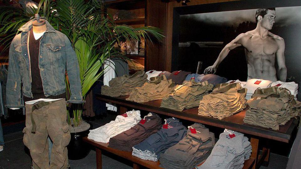 An interior shot of the Abercrombie & Fitch store on 5th Avenue in New York City, taken at its opening event in 2005, showing piles of folded clothes, a mannequin dressed in a denim jacket and cargo trousers and with an advertising image of a shirtless model in the background