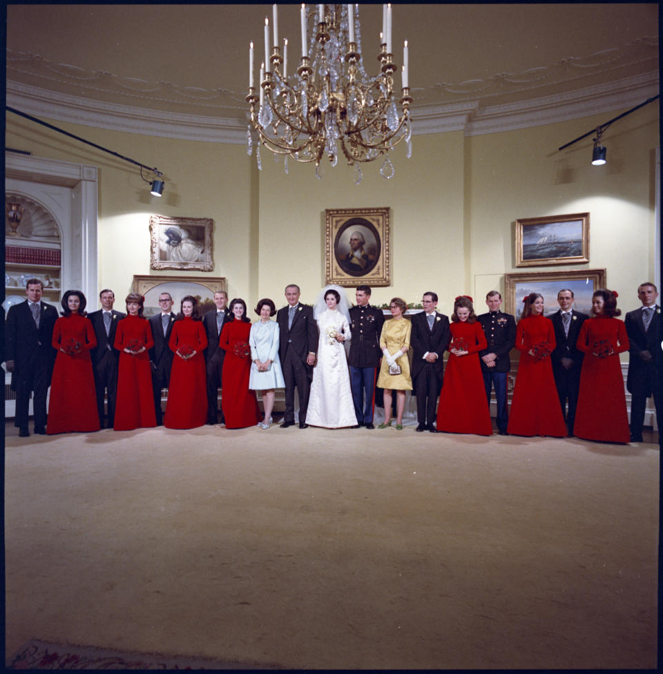 In this photograph by Frank Wolfe, President Lyndon B. Johnson and First Lady Lady Bird Johnson pose with their daughter, Lynda Bird Johnson, on her wedding day on Dec. 9, 1967. The Johnsons are joined by Lynda's groom, Marine Corps Capt. Charles S. Robb, her younger sister, Luci Baines Johnson, to the right of Mrs. Johnson, and the wedding party in the Yellow Oval Room on the second floor of the White House. This photograph was captured following Lynda and Charles' ceremony in the East Room. Along with the bridal ensemble, Geoffrey Beene designed the bridesmaids’ red velvet dresses, which were inspired by Francisco Goya’s 1787-1788 painting "Manuel Osorio Manrique de Zúñiga" and reflected the medieval aesthetic popularized by the 1967 film Camelot.