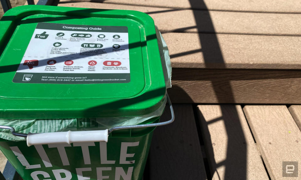 A green, square compost bin sits on wooden steps.  Instructions on what foods can go into the bucket are marked on the lid. 