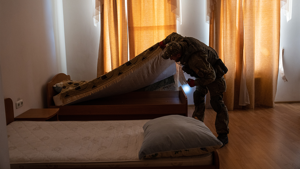 A Ukrainian soldier checks for IEDs under a mattress at Kupiansk, Kharkiv Oblast. The light is low.