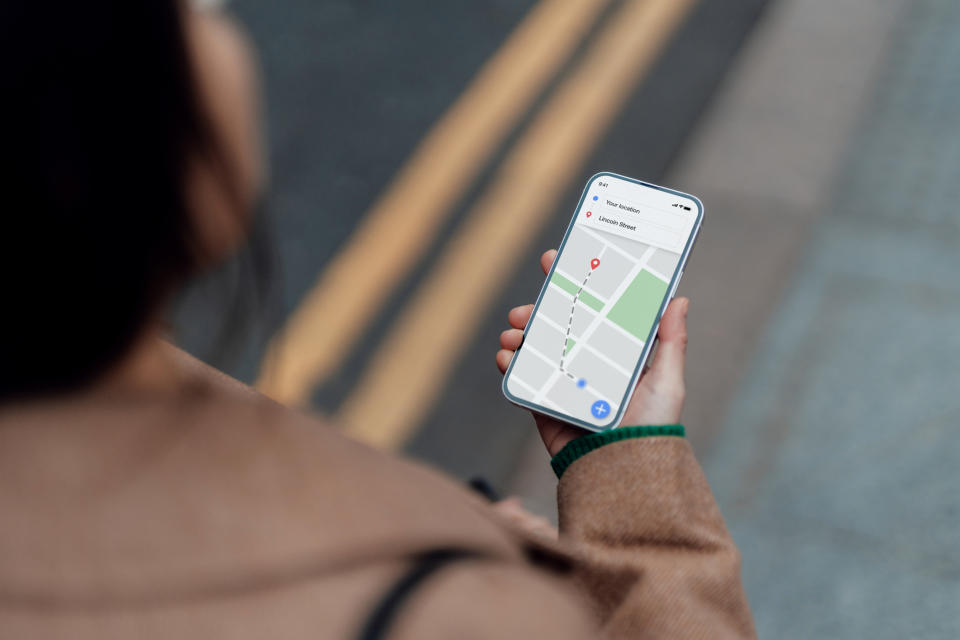 A person is holding a smartphone displaying a GPS navigation map with a red location pin. The individual appears to be walking along a street