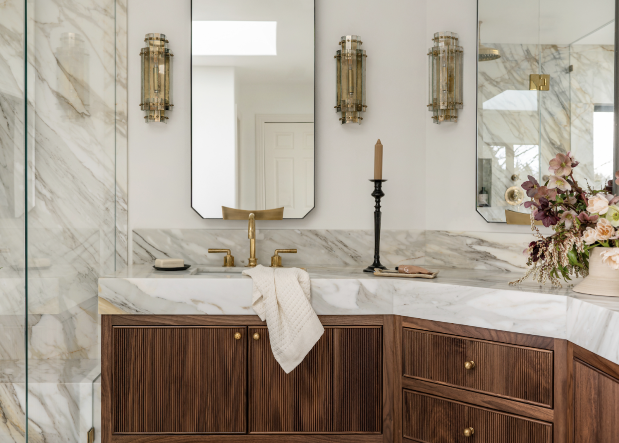  A beige bathroom with wooden cabinets, a marble counter top and luxe wall sconces. 