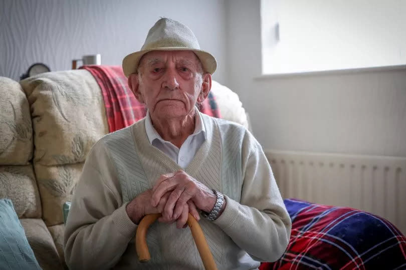 World War Two veteran, Norman Powell, 98, pictured at home in Toton, Nottingham. He is wearing a sweater and a white shirt and is seated and holding his walking stick.